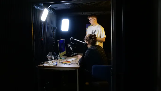 Two young men in a dark recording studio with bright lights, one sitting at a desk with a microphone and computer, the other standing beside him.