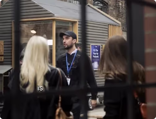 Man with a cap and lanyard speaking outside a small pop-up booth while two women face him from behind.