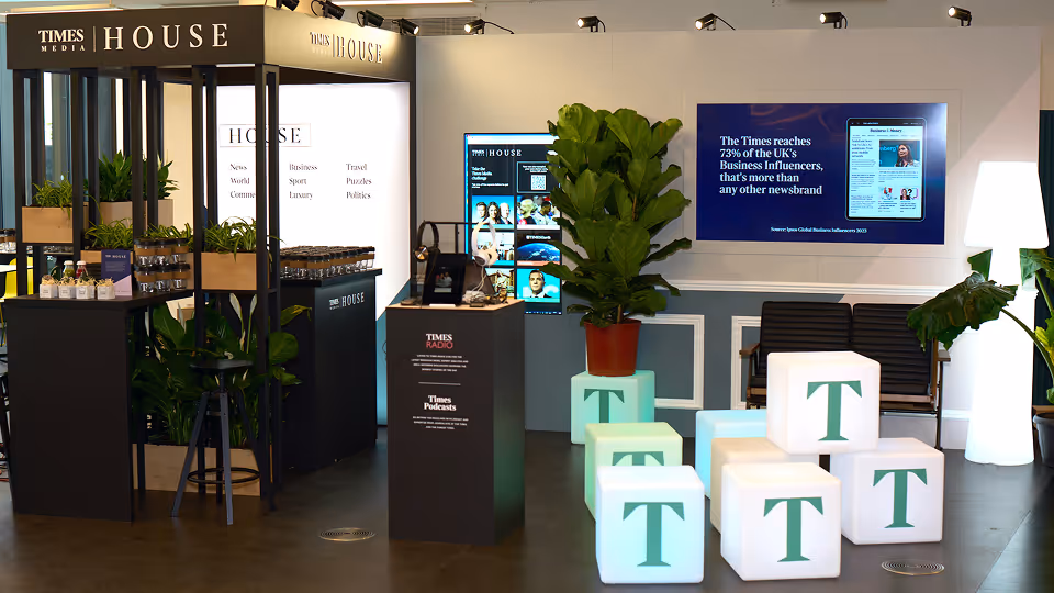 Modern Times House exhibition booth featuring illuminated cubes with 'T', plants, promotional displays, and digital screens in a styled room.