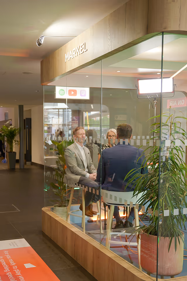 Three people seated and engaged in a discussion inside a glass-walled podcast studio labeled 'MARKEL' with professional lighting and plants visible.