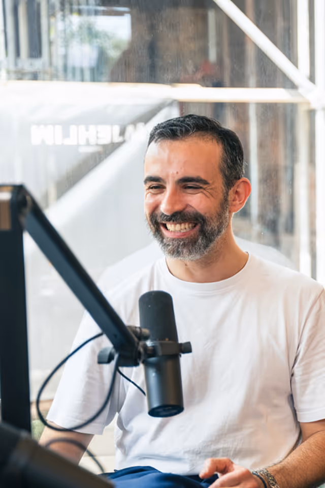 Smiling man with beard speaking into a microphone in a bright studio.