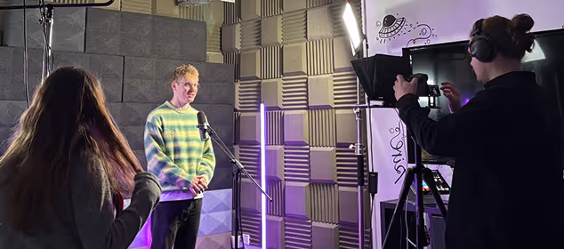 Man in striped sweater stands before microphone in soundproof room, filmed by cameraman with headphones on.