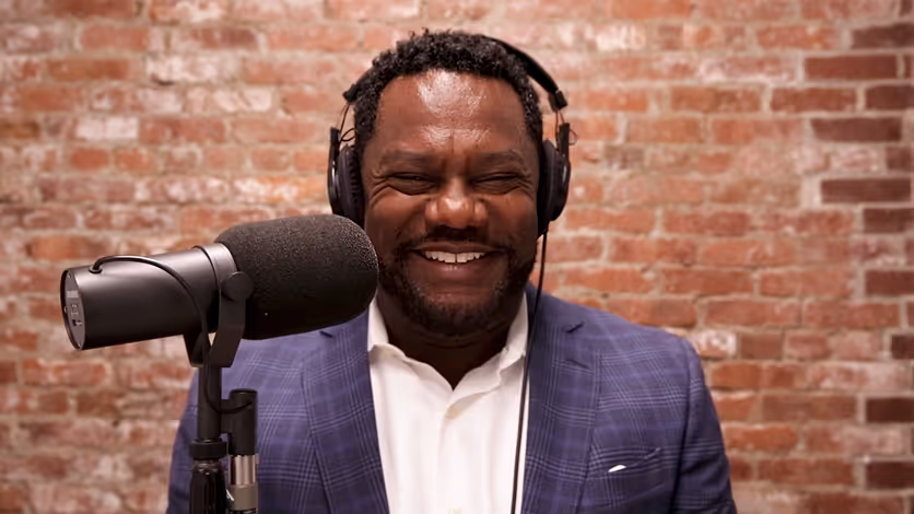 Smiling man wearing headphones and a blue suit speaking into a microphone with a brick wall background.