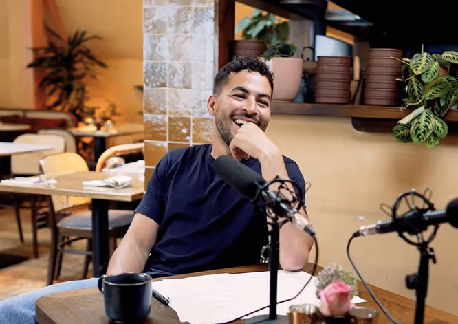 Smiling man with curly hair sitting at a table with microphones in a cozy cafe setting.
