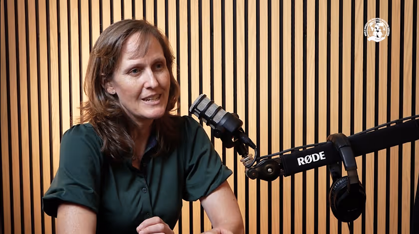 Woman speaking into a condenser microphone in a recording studio with wood panel background.