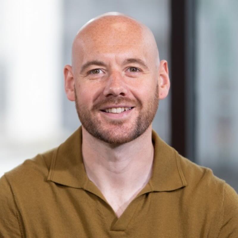 Shaun Staff from Azets. Smiling bald man with beard wearing a brown collared shirt against a blurred background.