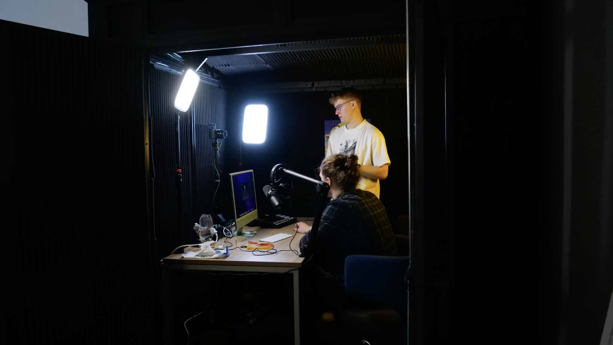 Two people working on video setup in dimly lit room with computer, microphone, and bright LED lights.