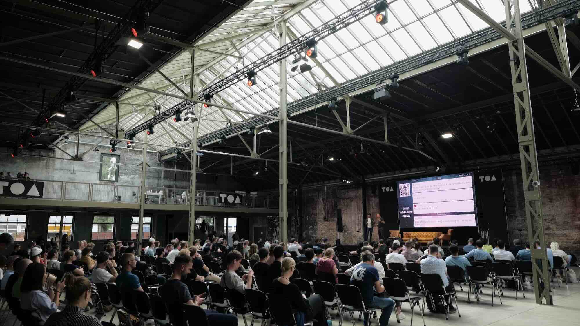 Audience seated in a large industrial-style hall watching a presentation on a big screen.