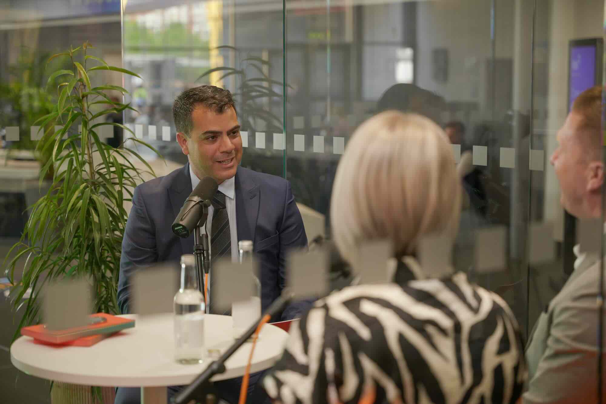 Man in suit speaking into a microphone across a table from two people in an office setting.