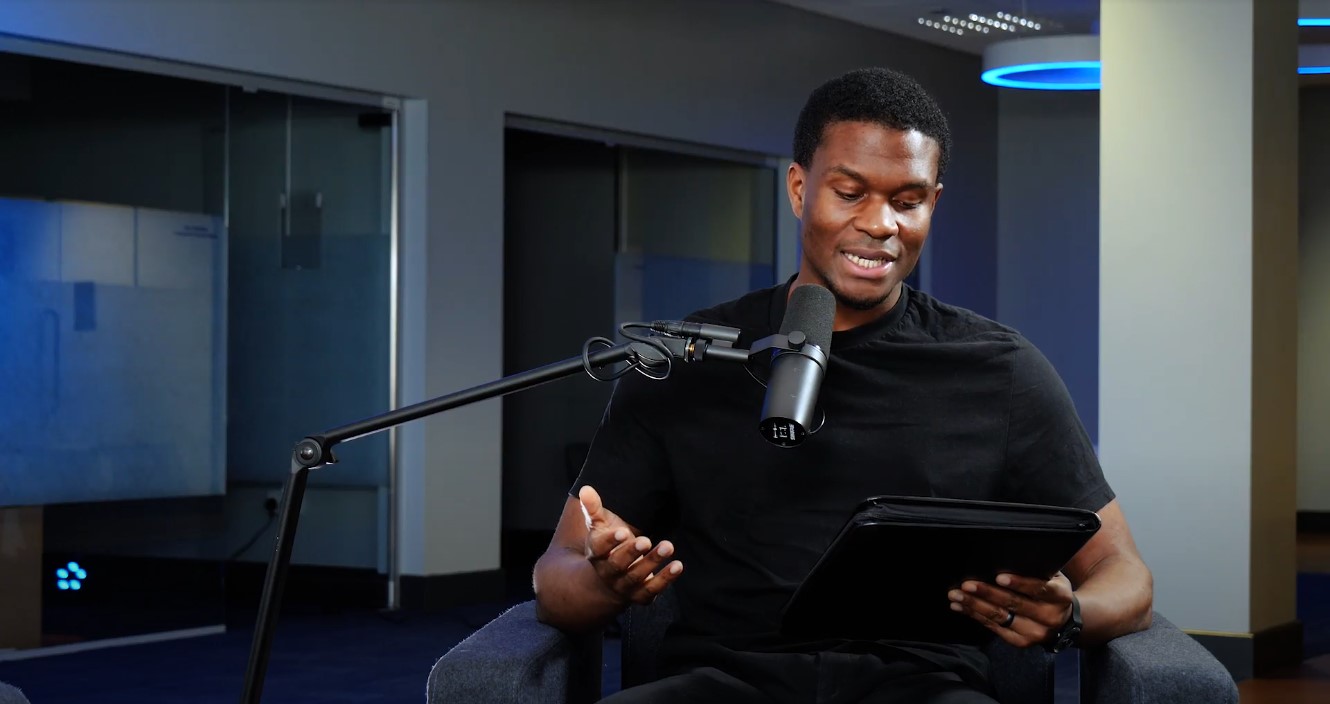 Man sitting in a chair holding a tablet and speaking into a microphone on a boom arm.