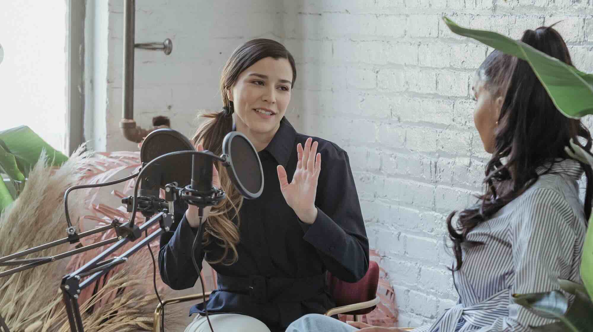 Two people chatting sat down in a podcast against a white wall.