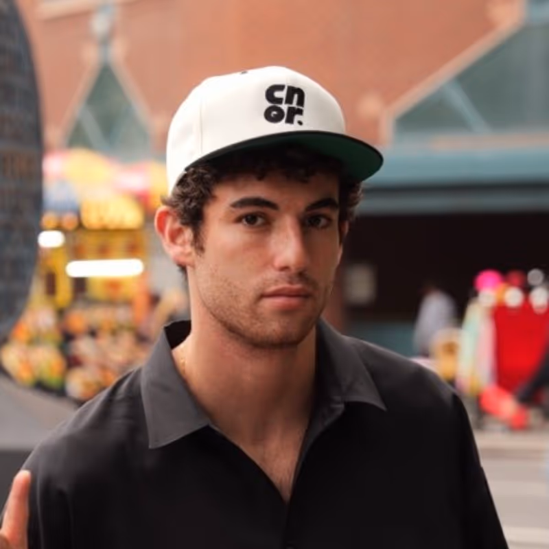 Young man with curly hair wearing a white cap and black shirt looking at the camera in an urban setting.