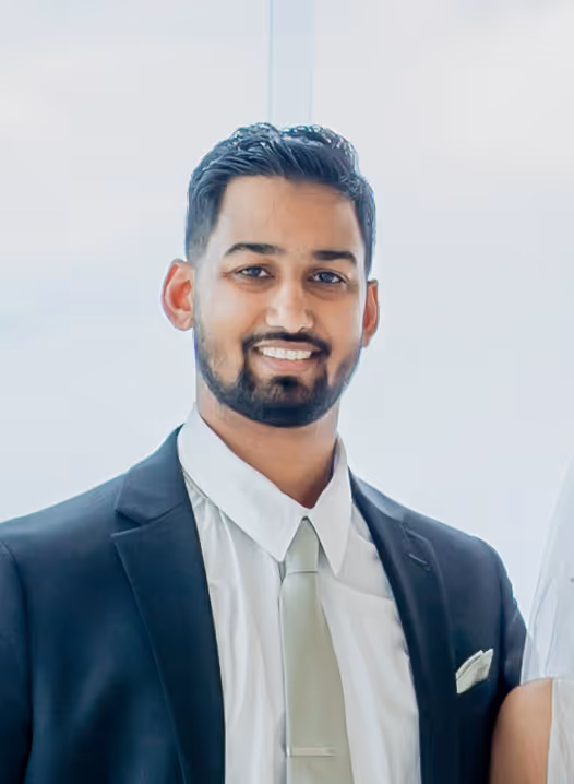 Smiling man with a trimmed beard wearing a black suit, white shirt, and light-colored tie.