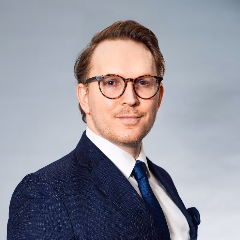 Man with light brown hair and tortoiseshell glasses wearing a navy suit, white shirt, and blue tie against a gray background.