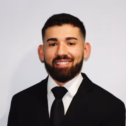 Portrait of a smiling man with short dark hair and beard wearing a black suit, white shirt, and black tie against a plain light background.