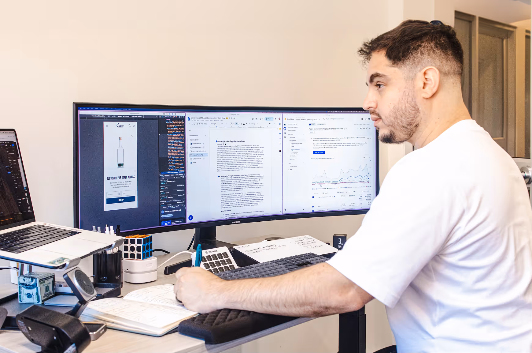 Man in white shirt working at a desk with an ultra-wide monitor displaying code, a document, and analytics, while writing in a notebook.