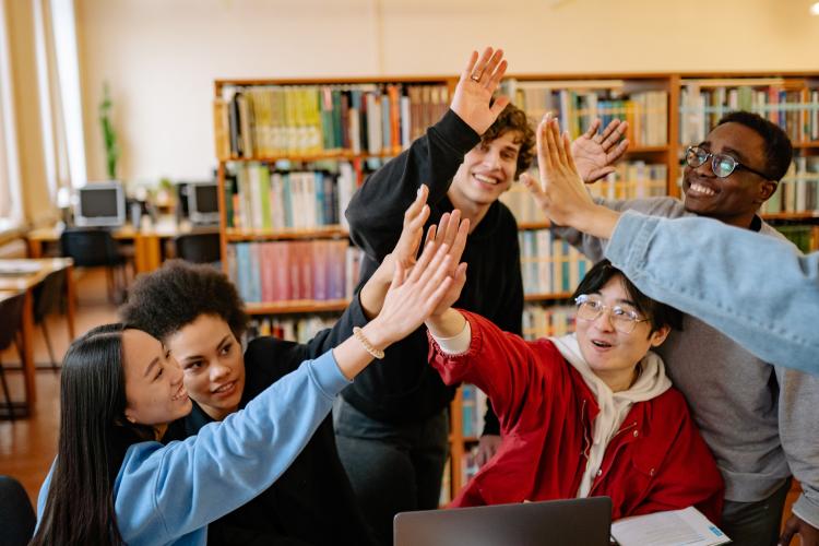 A group of high school students enjoying their academic year in America smile and high five. Bright library shelves are visible in the background.