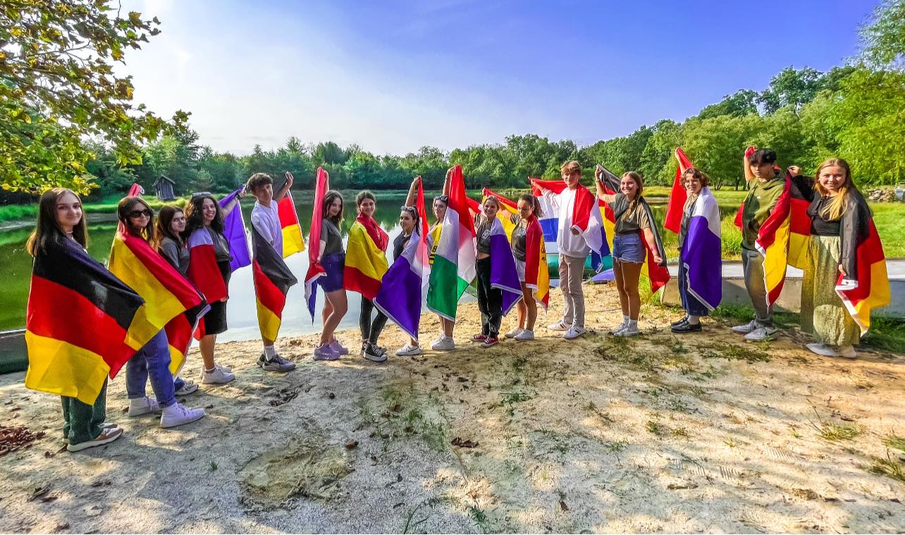 A group of young foreign exchange students draped in their home countries' flags poses for a picture in front of a lake
