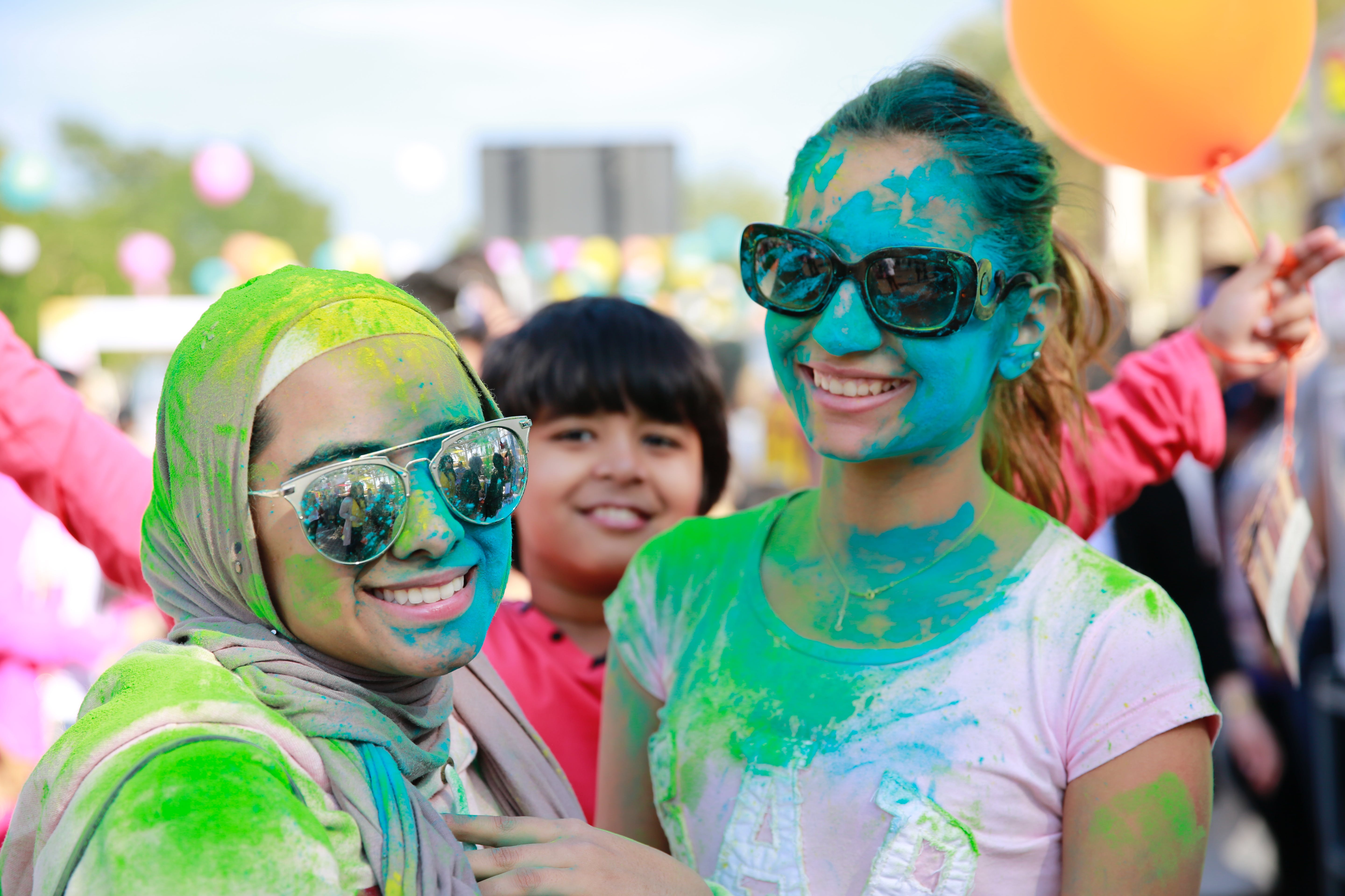 Two girls wearing sunglasses and covered in colorful dust smile for the camera as a young boy photobombs their picture.