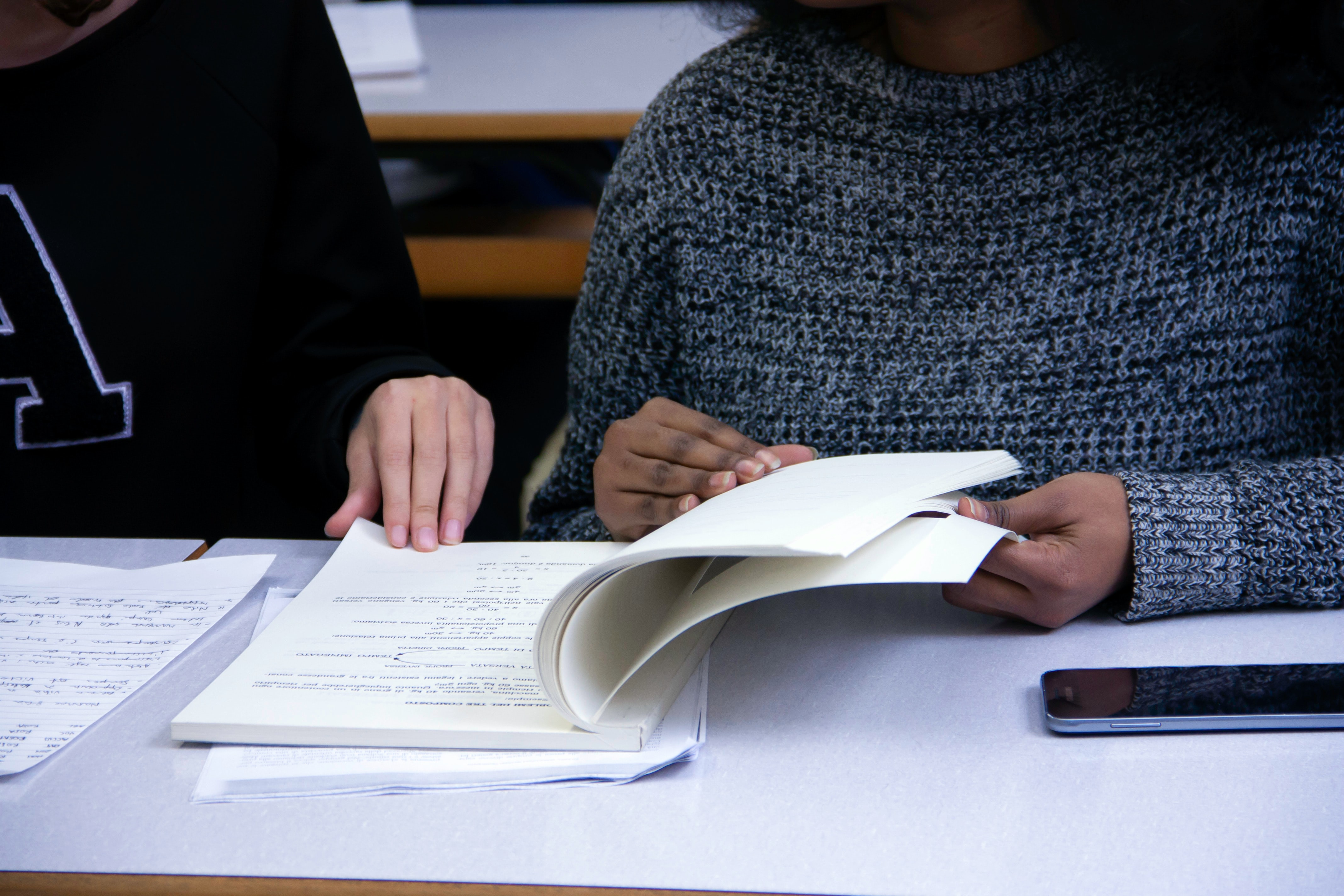 Two individuals sit side by side flipping through a textbook. Only their hands and upper bodies are visible, and they are wearing sweaters and college-lettered shirts, as they appear to be in an educational setting.