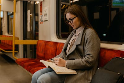 A young woman in glasses, jeans, and a grey overcoat sits alone in a cushioned subway line or train, writing in a notebook.