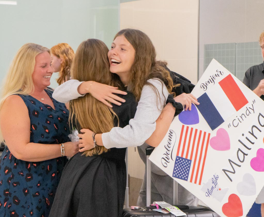 A foreign exchange student smiles while hugging her host family as they greet her with signs at the airport.