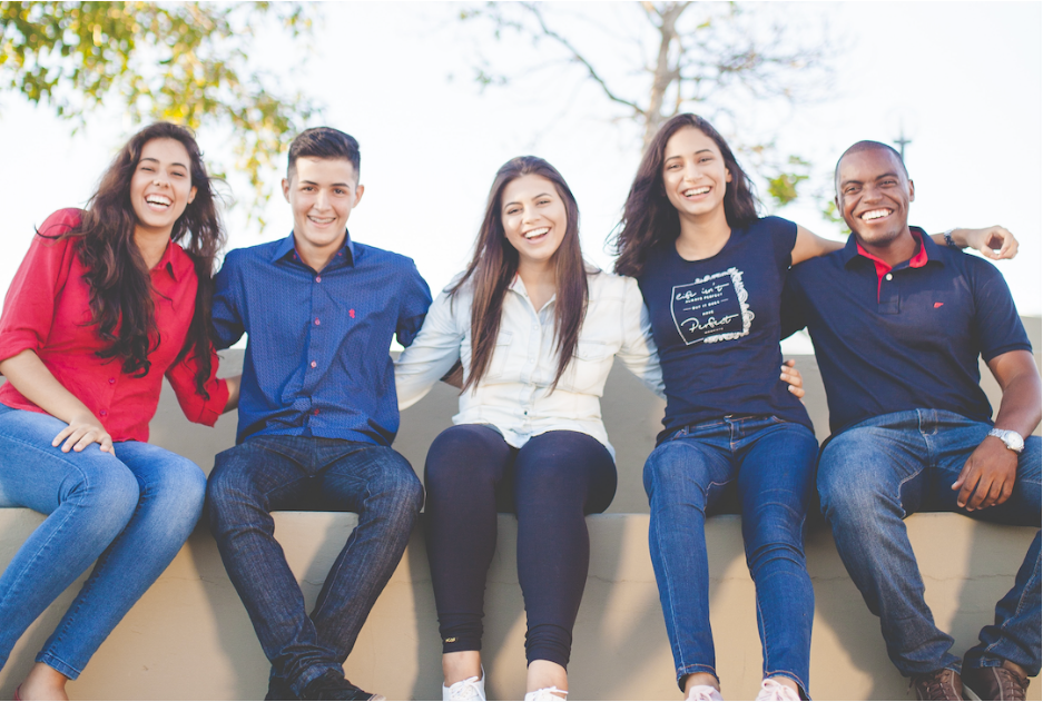 A group of young adults of varying genders and races sit on a low wall, smiling and with arms around each other's shoulders