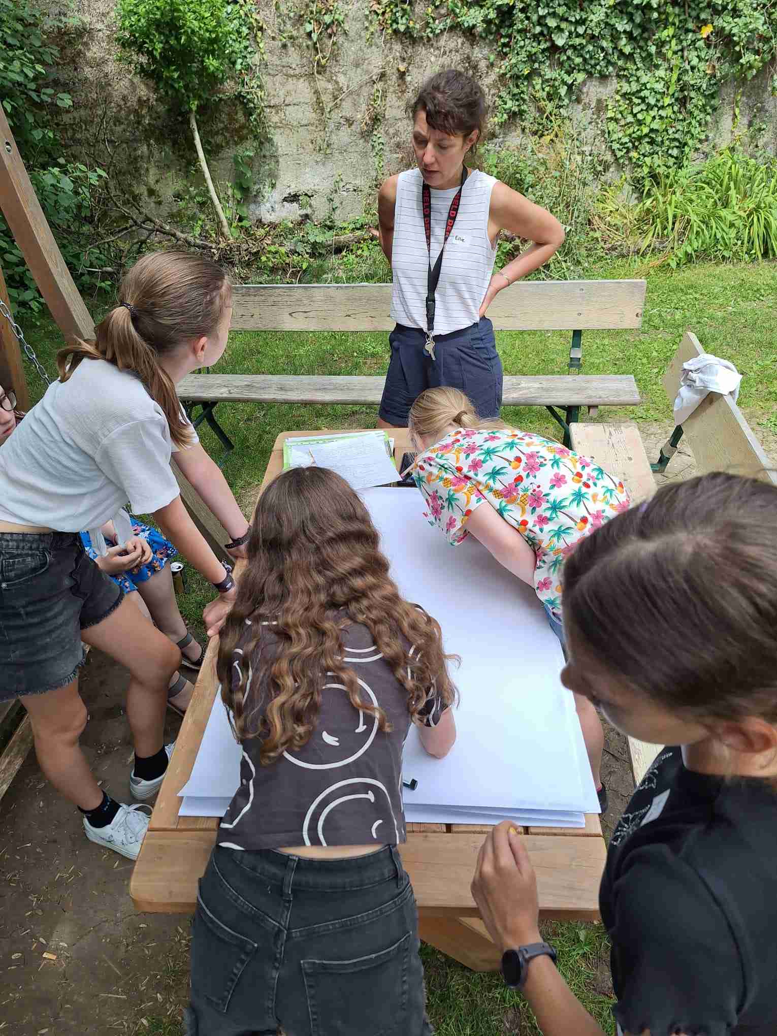 Eine erwachsene Frau steht an einem Holztisch im Freien und berät vier Mädchen, die sich über einen großen weißen Bogen Papier beugen.