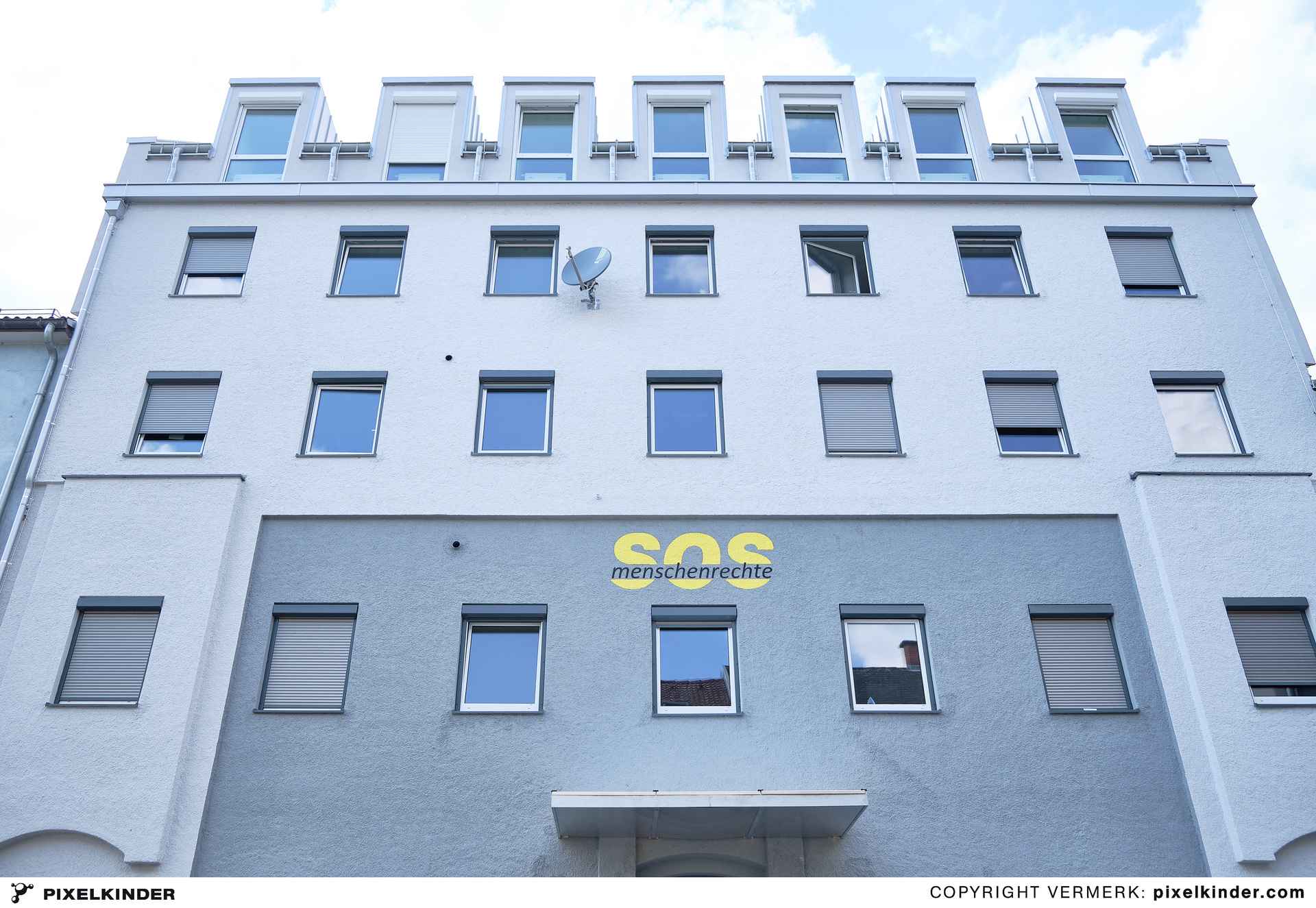 Fassade eines weißen mehrstöckigen Gebäudes mit mehreren Fenstern und dem gelben Schriftzug 'SOS Menschenrechte' auf grauem Hintergrund.