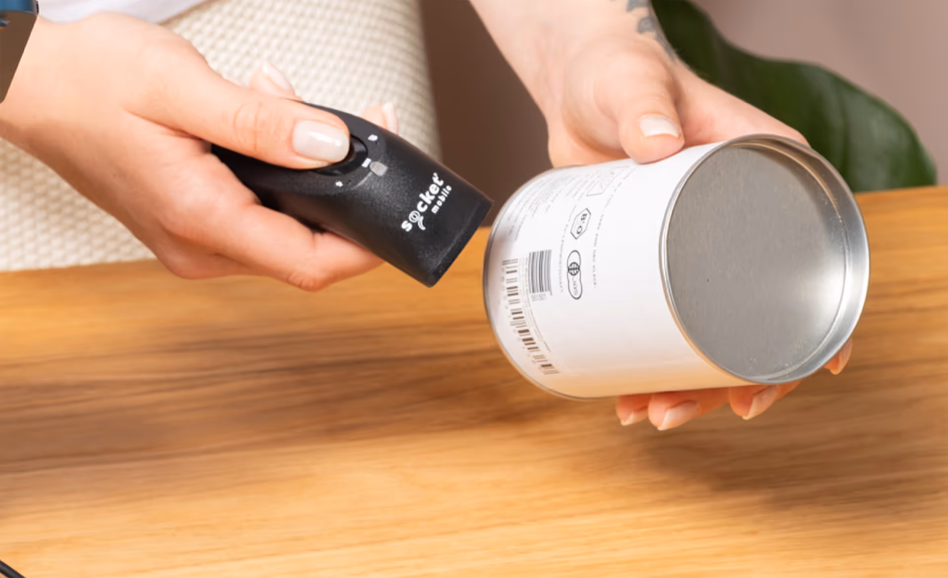 Eine Hand hält einen schwarzen Scanner über eine Weißblechdose mit Barcode auf einem Holztisch.