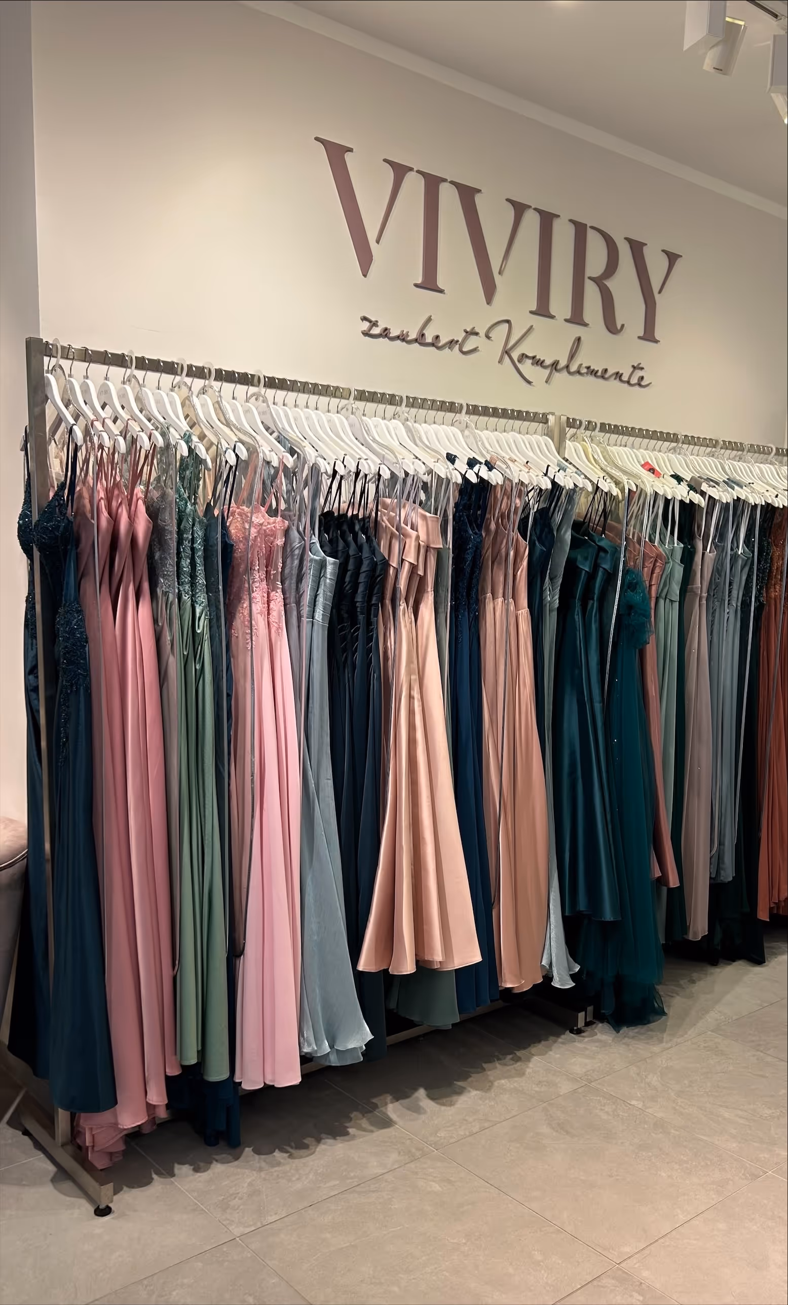 Kleider in verschiedenen Pastellfarben hängen ordentlich auf Kleiderbügeln in einem eleganten Bekleidungsgeschäft vor einer Wand mit dem Schriftzug ‚VIVIRY Zaubert Komplimente‘.