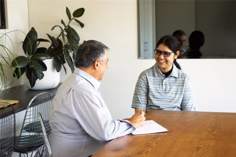 A male sitting at a table, talking with a female who is looking at him and smiling, the male has a notepad on the table.