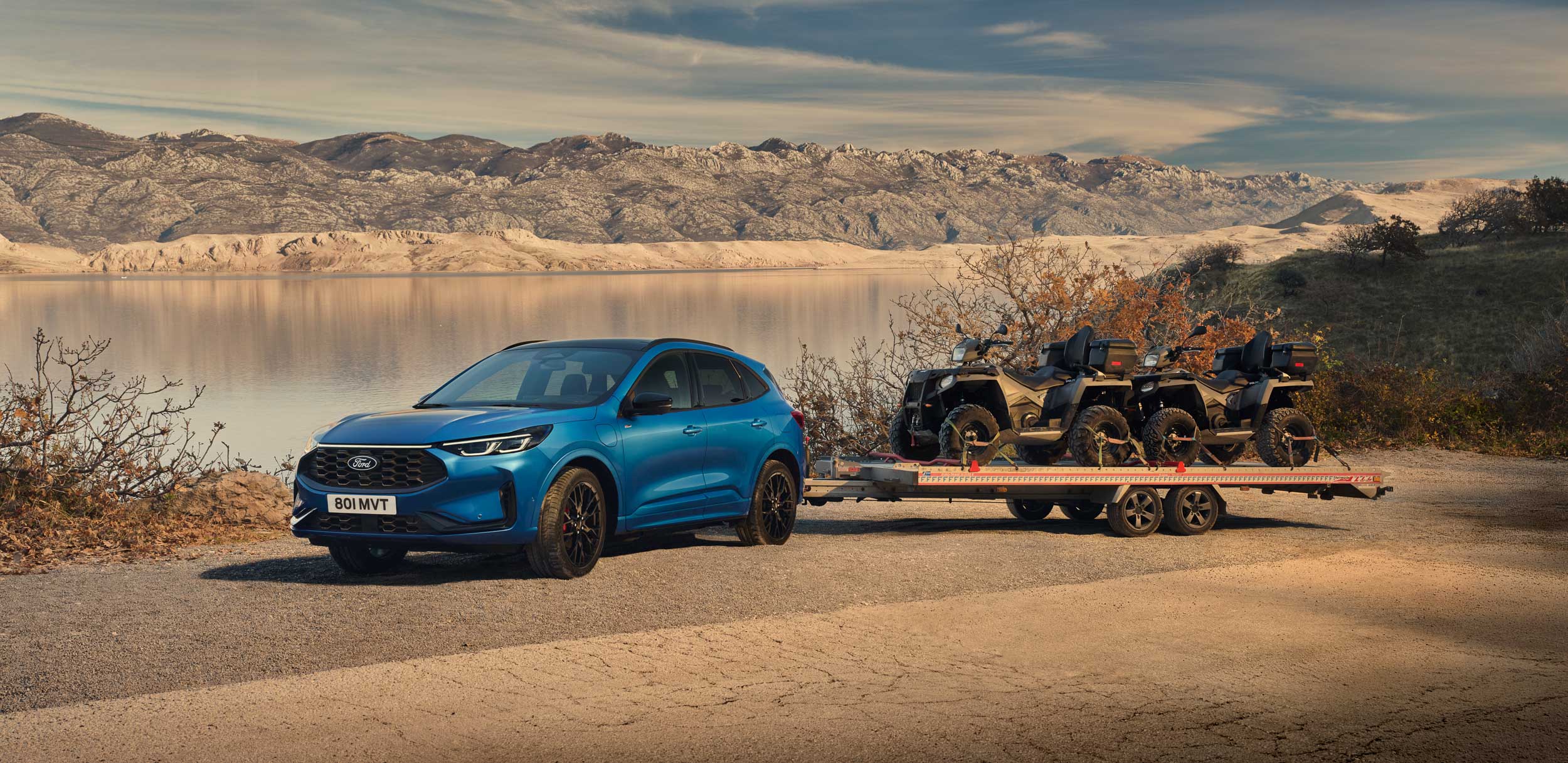 Blue Ford Kuga towing quad bikes beside a mountain lake.
