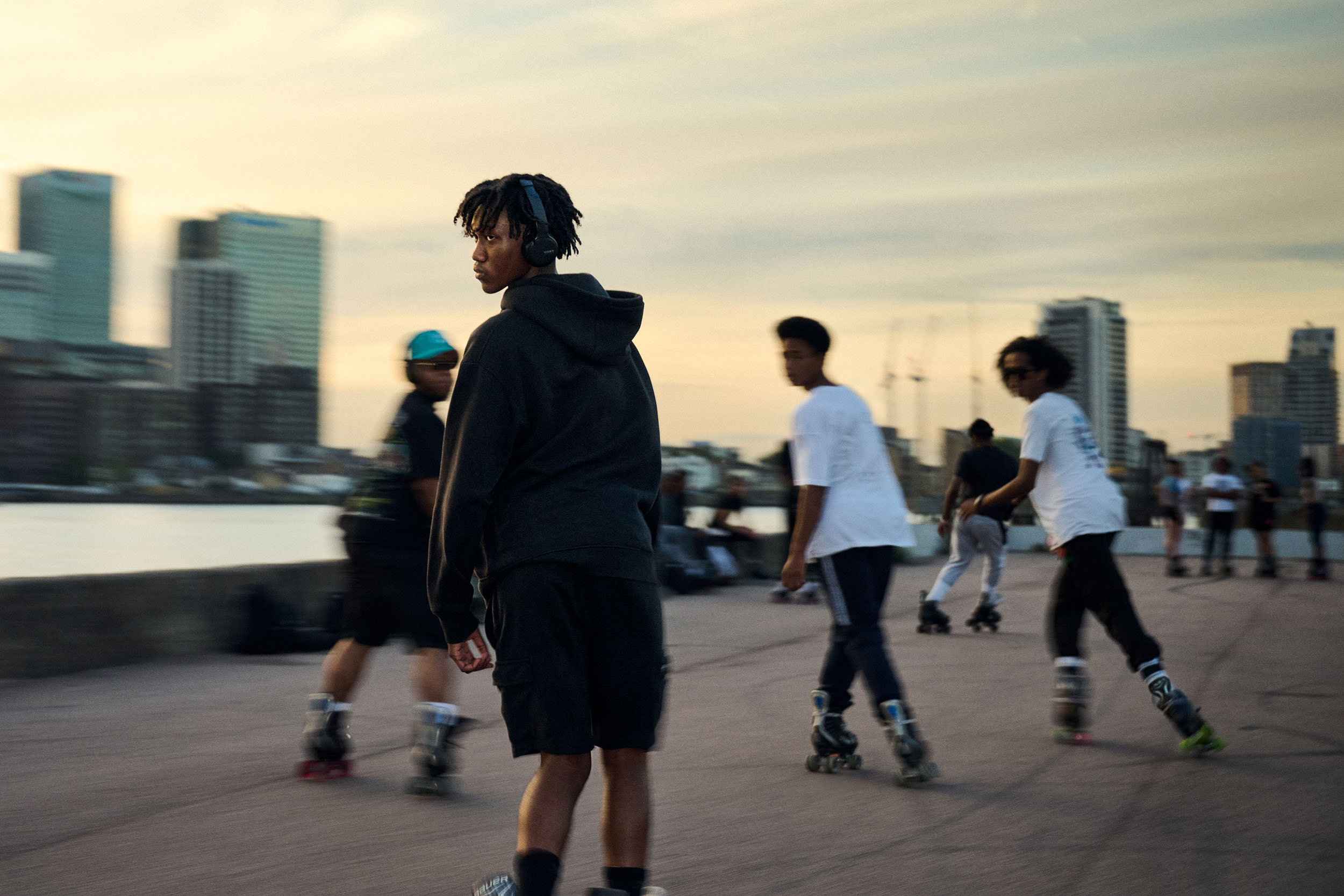 A skater looks back over his shoulder as the group circles, Canary Wharf behind at sunset.