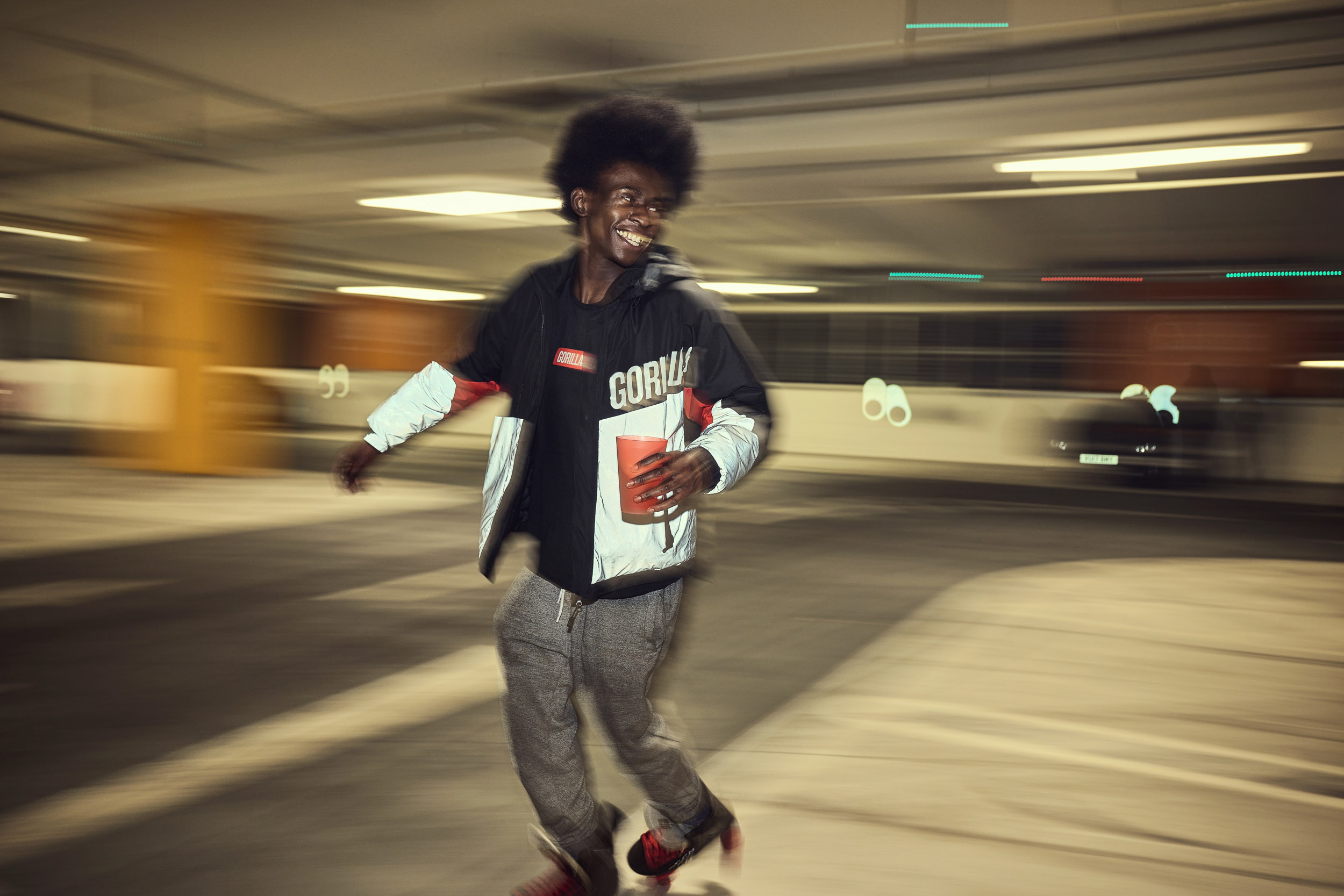 A skater rolls past with a drink in hand, his reflective jacket catching the car park lights.