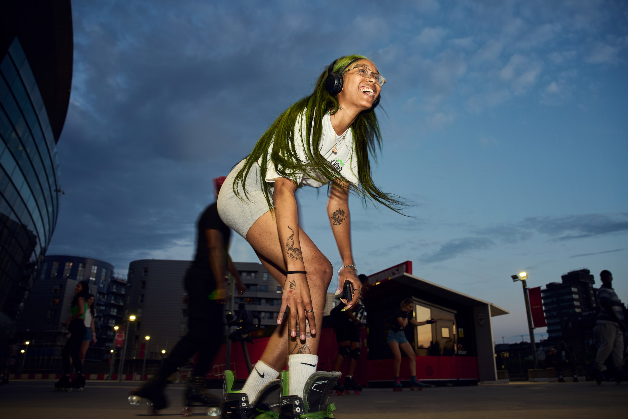 A skater with green hair laughs mid roll, the flats and the stadium behind her.
