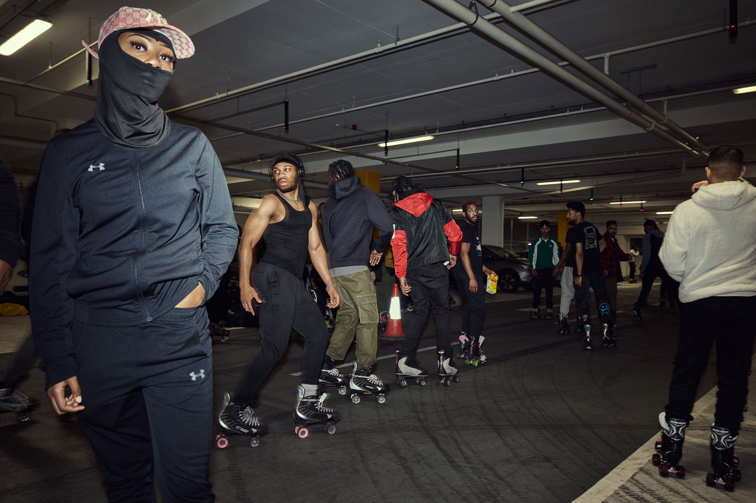 A skater in a tracksuit and balaclava stands in front of a floor crowded with skaters.