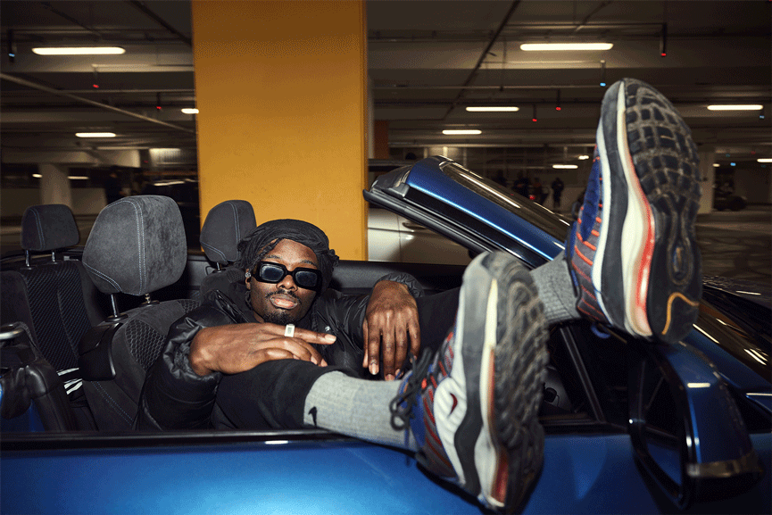 Skater in sunglasses reclining in an open-top car with feet up on the windscreen, London car park.