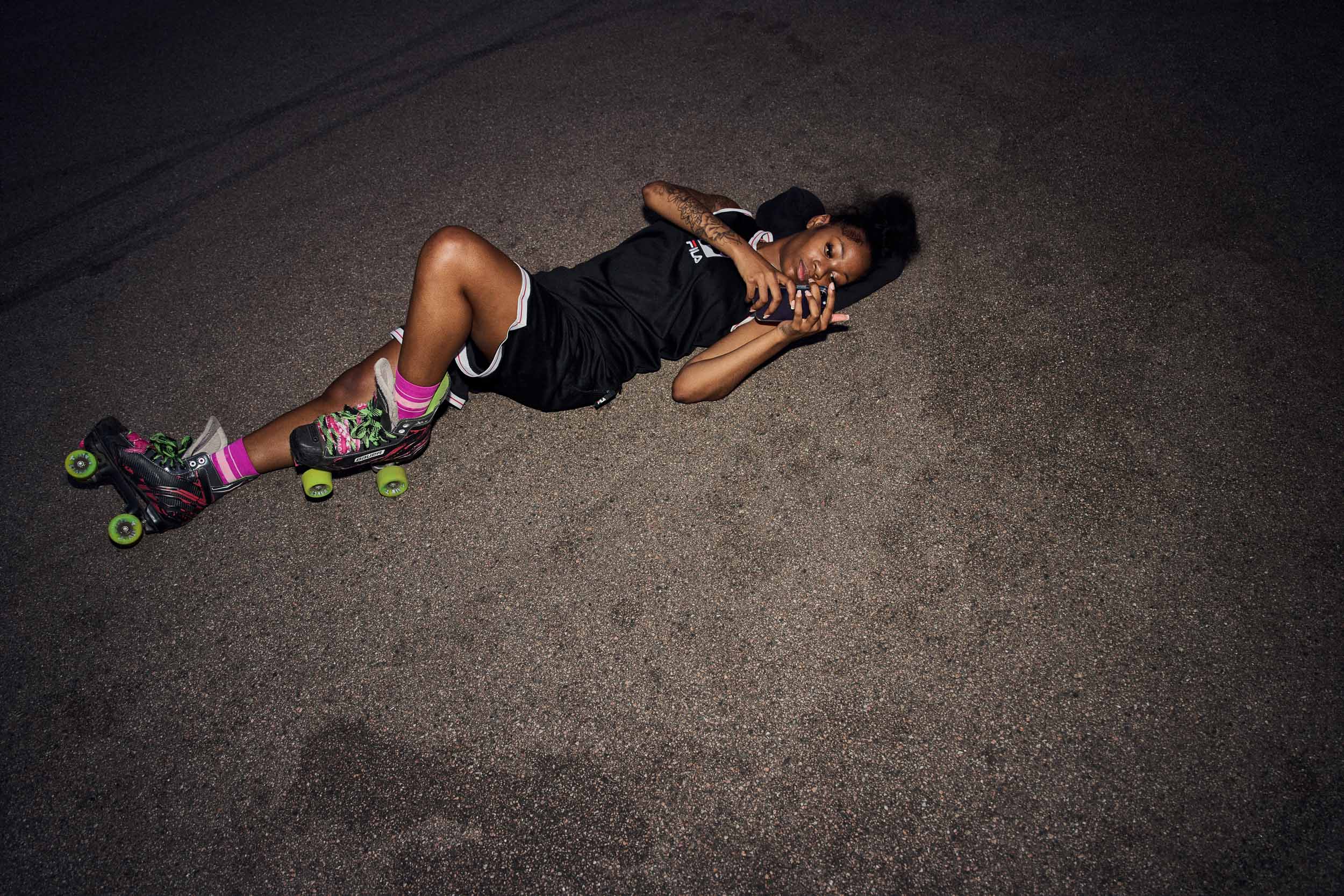 A skater lies back on the tarmac with her phone, skates still on, late in the night.
