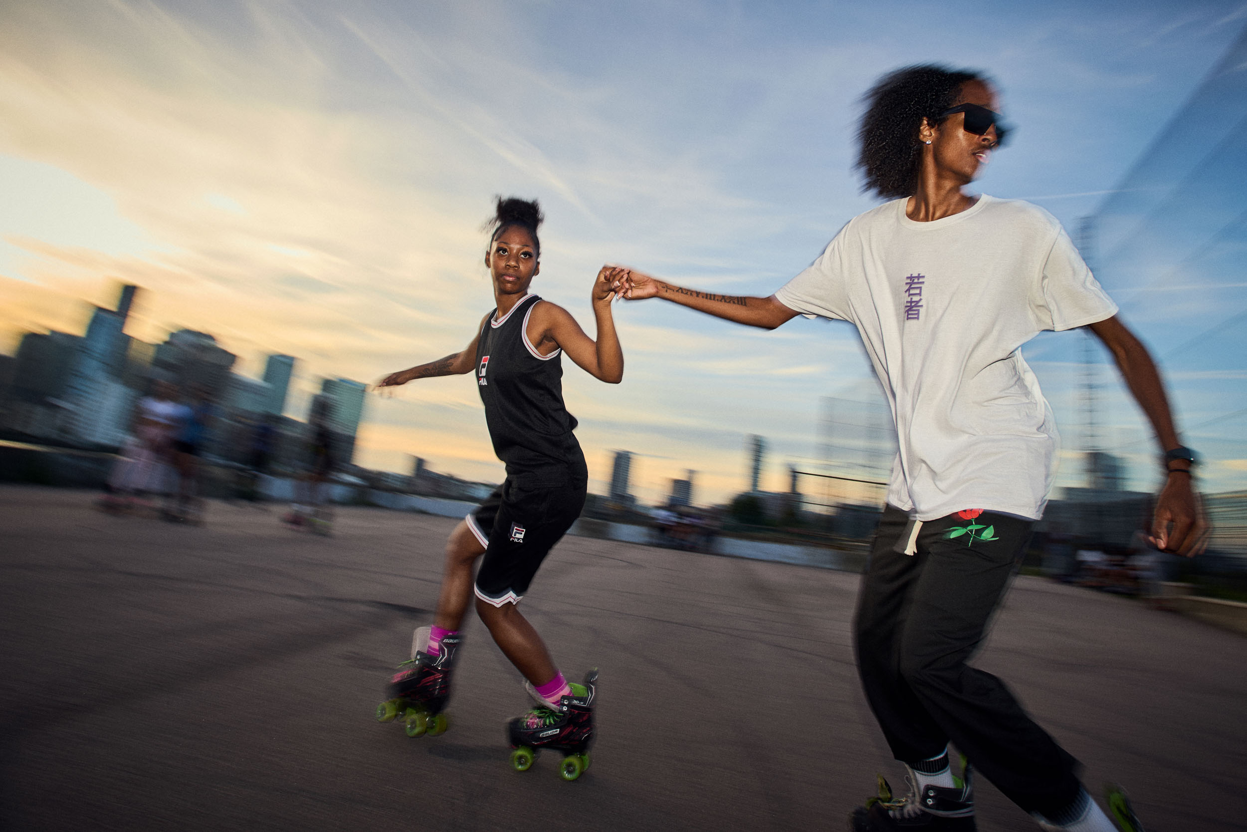 Two skaters hold hands as they roll along the riverside at sunset.