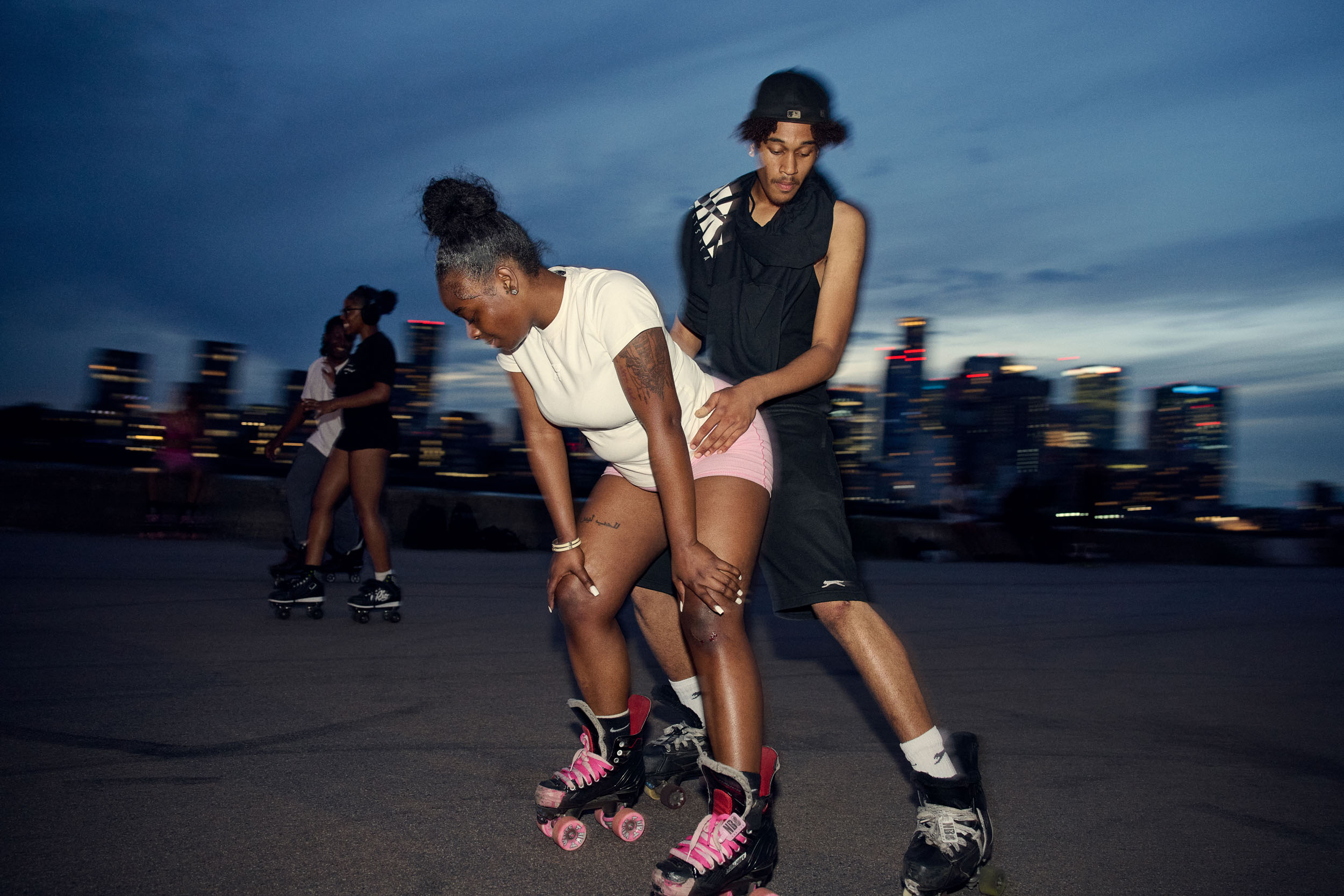 Two skaters move together in the dark with the lights of Canary Wharf behind them.