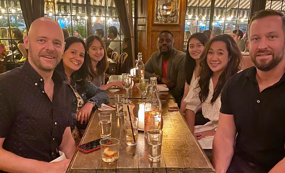 Seven people sitting at a wooden table in a warmly lit restaurant, smiling at the camera.