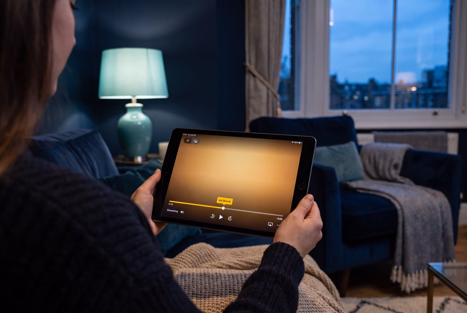 Viewer on a couch streaming video on a tablet, the player showing an Ad Break marker on the progress bar (AI-generated illustration)