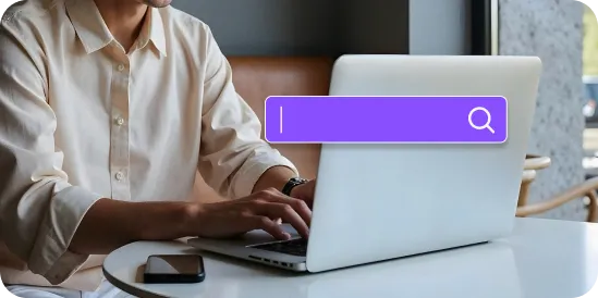 Person typing on a laptop at a table with a smartphone nearby and a search icon overlay.