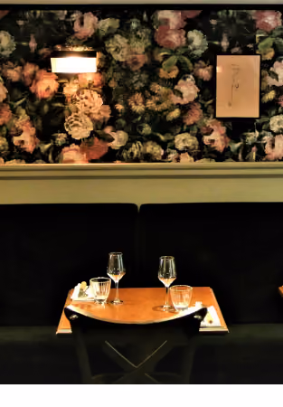 Restaurant table set for two with glasses and napkins, in front of a wall with floral wallpaper and a black bench.