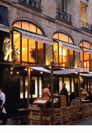 The facade of a Parisian restaurant lit up with large rounded windows, where customers dine on the terrace under a dark sky.