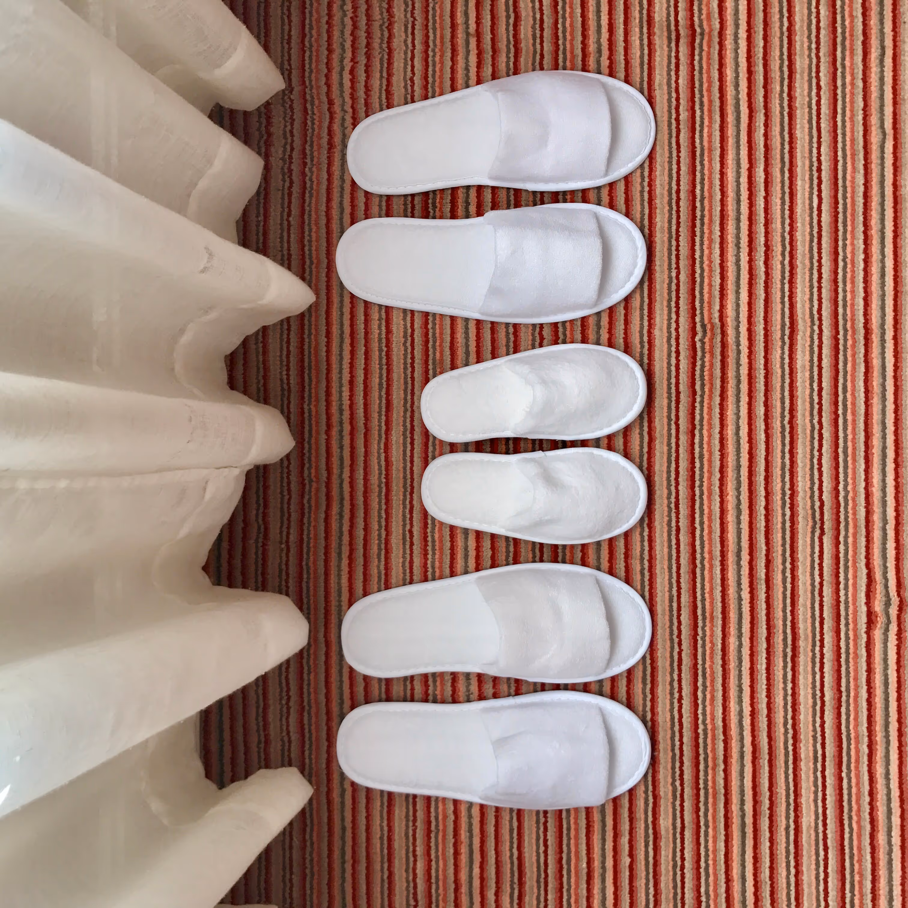 White slippers lined up on a red striped rug near curtains in an elegant interior space.