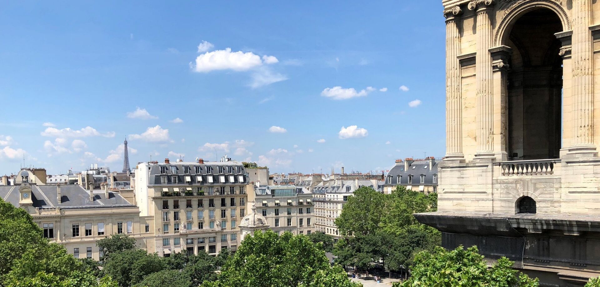 Vue panoramique sur Paris avec la tour Eiffel, les toits haussmanniens et l’Arc de Triomphe sous un ciel bleu.