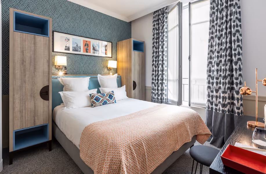 Chambre d'hôtel lumineuse avec grand lit double, deux armoires en bois, lampes murales, rideaux à motifs noirs et blancs, et bureau avec lampe et plateau rouge.