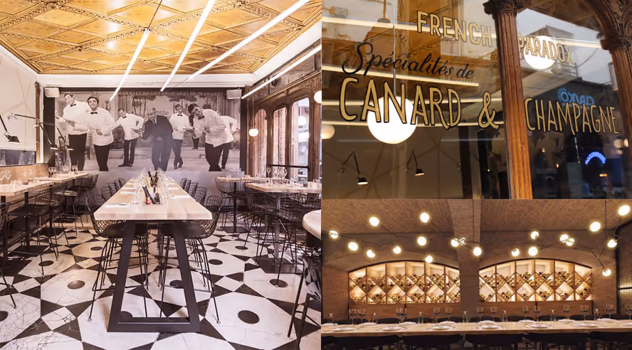 Interior of an elegant restaurant with a long table set for dining, a large mural depicting waiters in white uniforms, wooden walls and black and white geometric tile flooring, a display case with the inscription "French Paradox Specialties of Duck & Champagne," and a corner with wine racks illuminated by round hanging lamps.